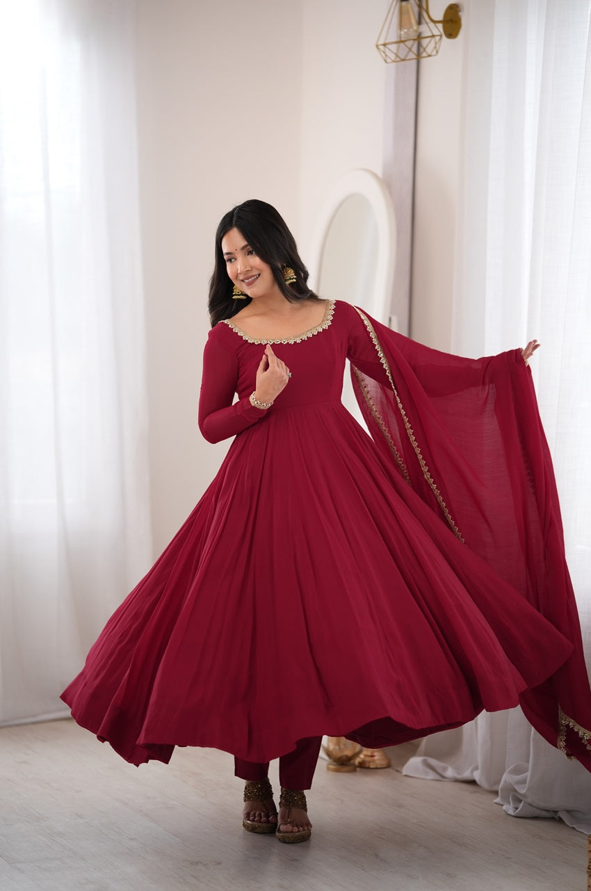 A woman modeling a long-sleeved maroon red Anarkali gown with a matching dupatta in a bright, elegant room with white curtains.