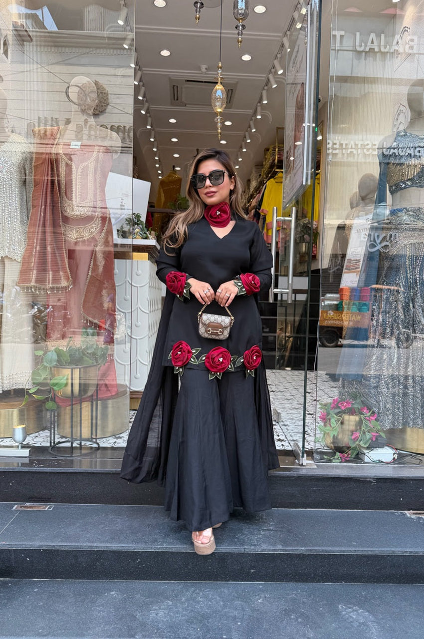A woman stands in front of a clothing boutique wearing a long black dress with red rose embellishments and red rose cuffs, accessorized with a red collar, sunglasses, and a small metallic handbag.
