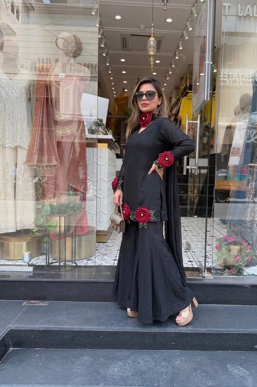 A woman models a black long-sleeve kurta with red rose embroidery, paired with a matching black dupatta, standing in front of a clothing boutique window.