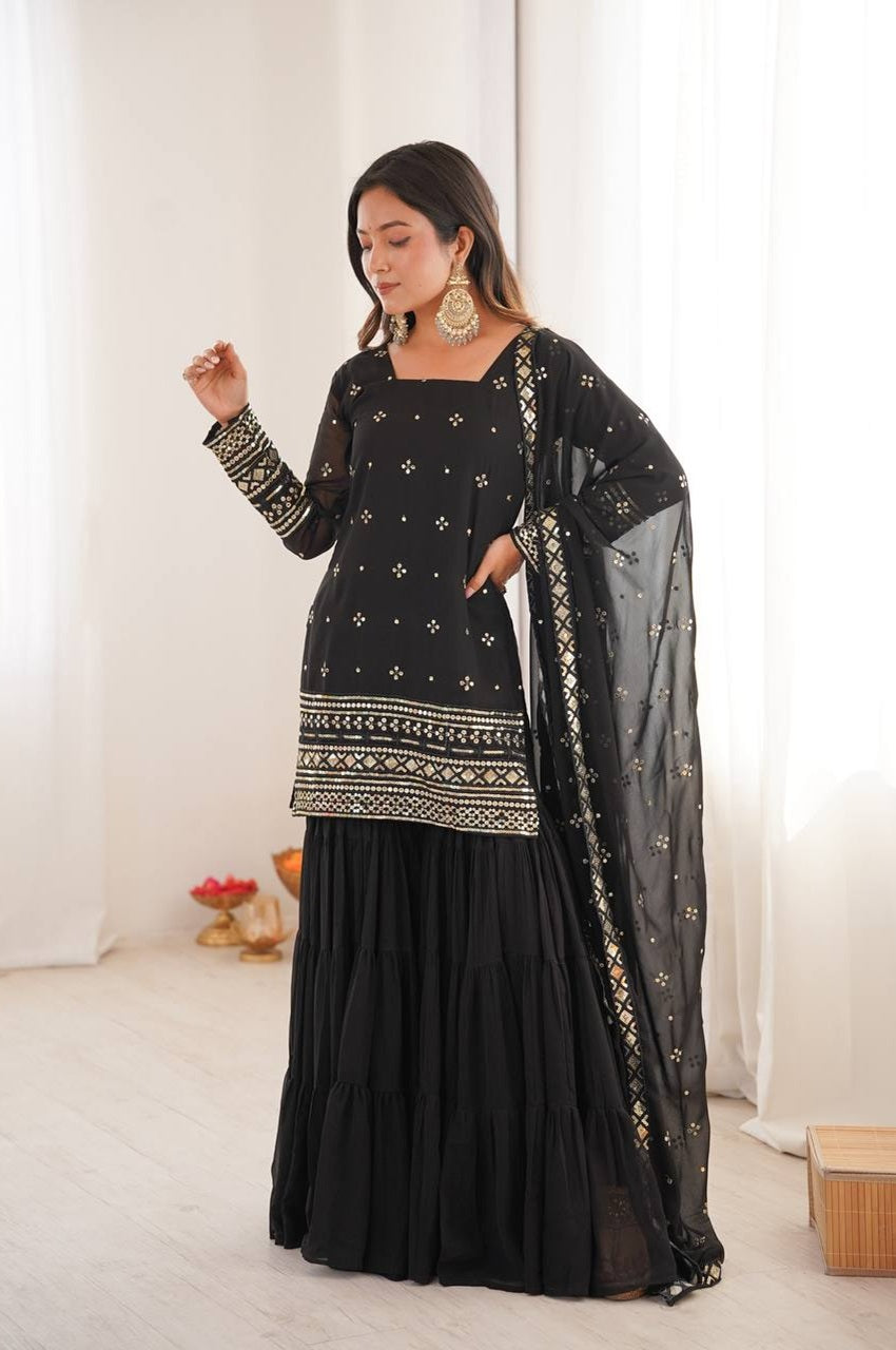 Woman wearing a black traditional outfit with intricate patterns in a bright room.