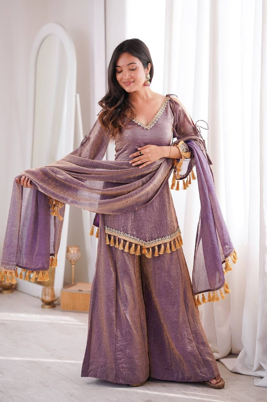 A woman models a traditional Indian outfit consisting of a lavender-purple velvet kurta with gold embroidery, matching wide-leg trousers, and a sheer purple dupatta with gold tassels, 