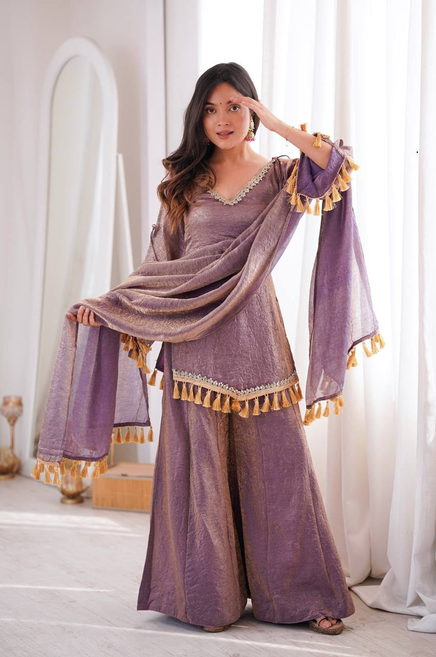 A woman models a traditional Indian outfit consisting of a lavender-purple kurta with wide legs, matching wide sleeves with gold tassel trim, and a coordinating dupatta draped over her shoulders. 