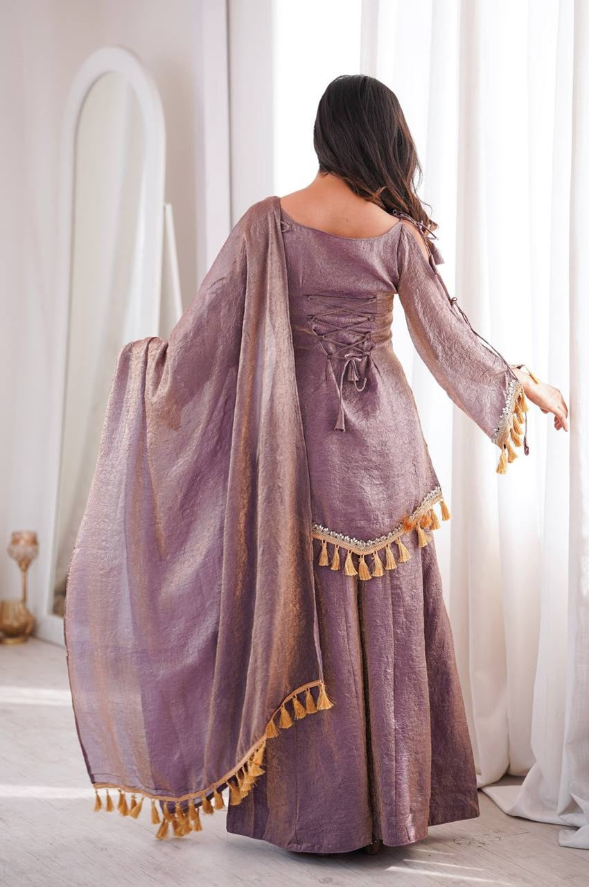 A woman wearing a lavender-purple traditional dress with a matching sheer dupatta and gold tassels, shown from the back in a bright, minimalist room with white curtains.
