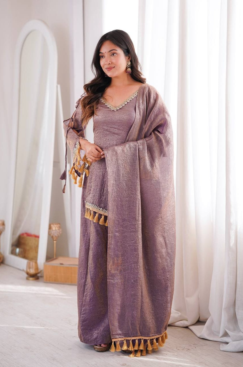A woman modeling a lavender-purple traditional outfit with gold tassels and silver embellishments, standing in a bright room with white curtains and a mirror.