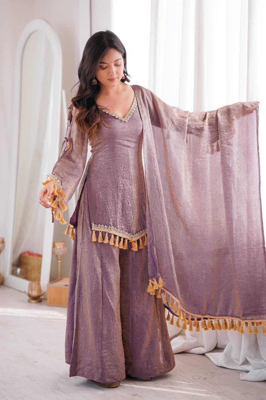A woman models a traditional Indian outfit consisting of a lavender-purple kurta with gold tassel trim, matching wide-leg trousers, and a sheer lavender dupatta with gold tassels,