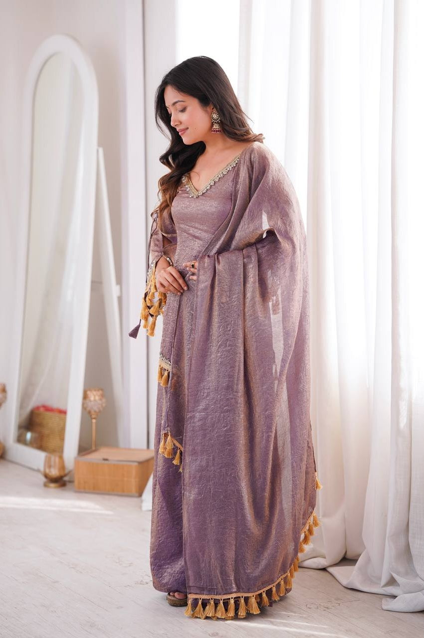 A woman wearing a lavender-purple traditional dress with gold tassel trim, standing in a bright room with white curtains and a mirror in the background.