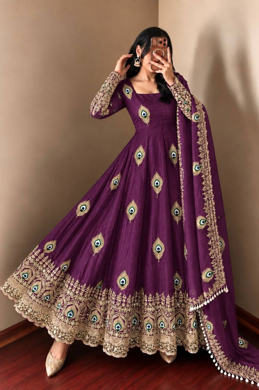 A woman wearing a purple Anarkali gown with gold and green peacock-eye embroidery and a matching dupatta, taking a mirror selfie in an indoor setting.