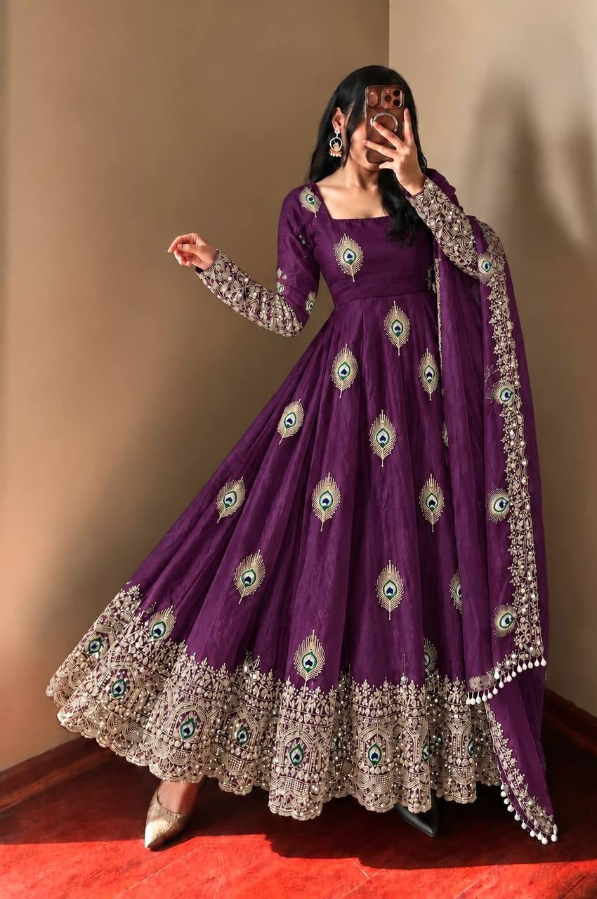 A woman modeling a deep purple Anarkali gown with intricate silver and gold embroidery, featuring peacock-eye motifs and a matching embroidered dupatta, set against a plain beige wall.