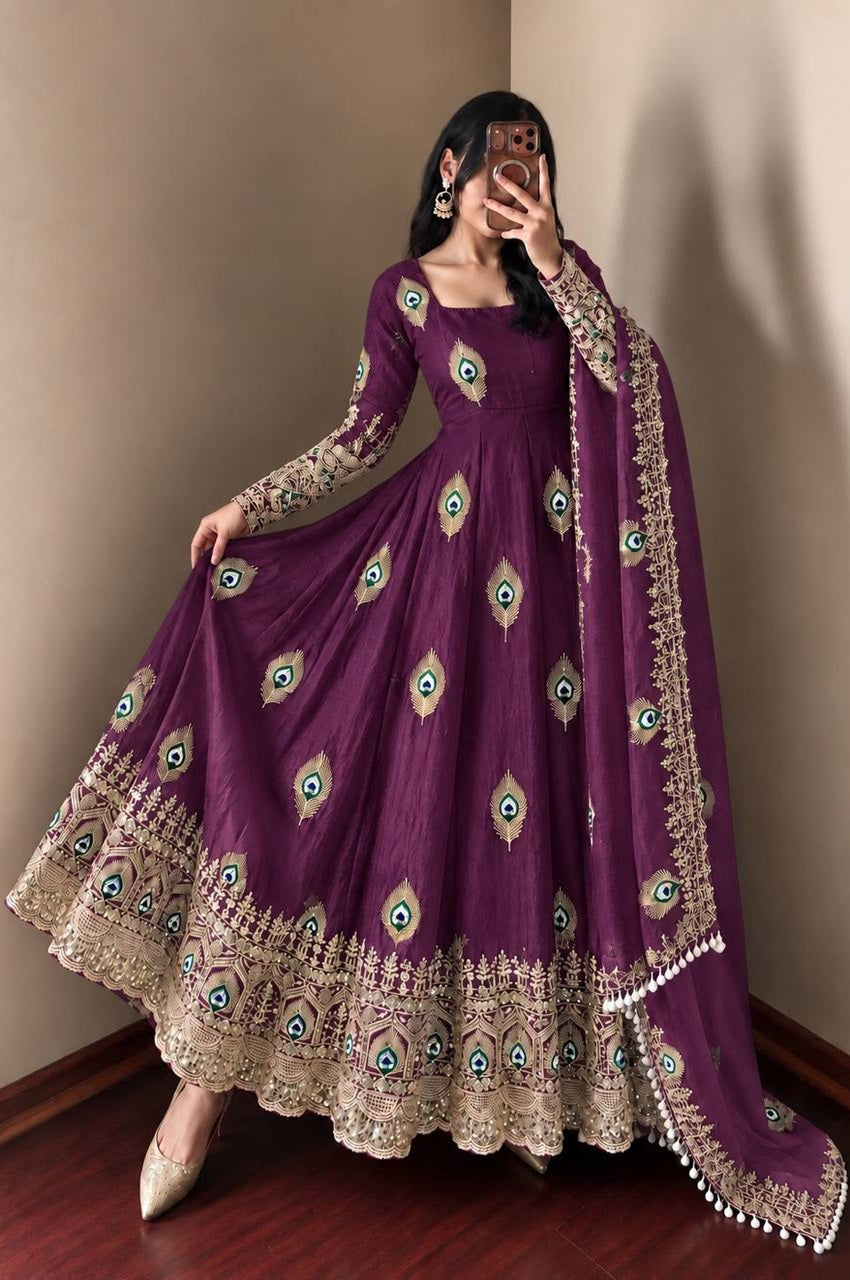 A woman models a deep purple Anarkali gown with gold and green peacock-eye embroidery and a matching dupatta, standing in a corner with a beige wall and dark wood floor.