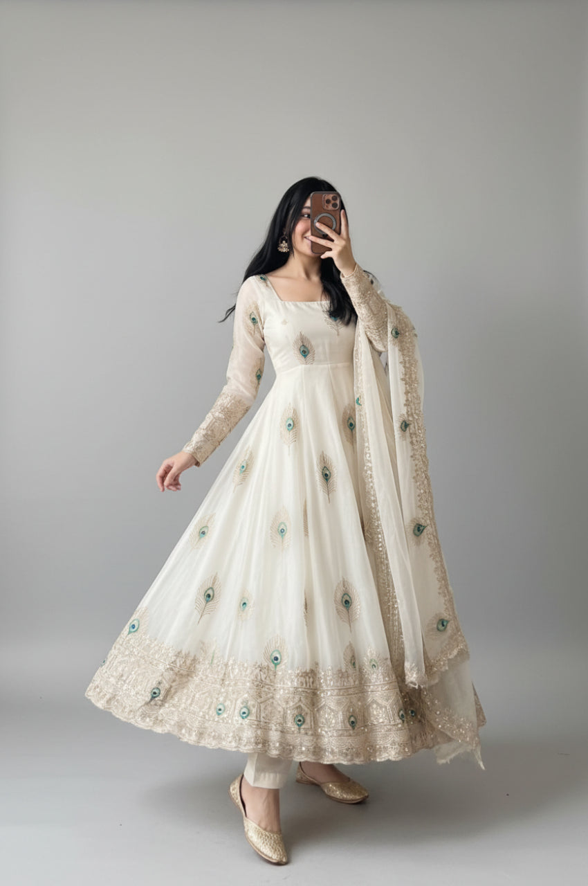 A woman takes a mirror selfie wearing a long-sleeved cream/off-white Anarkali gown with intricate gold embroidery and scattered teal green floral accents, paired with a matching sheer dupatta.