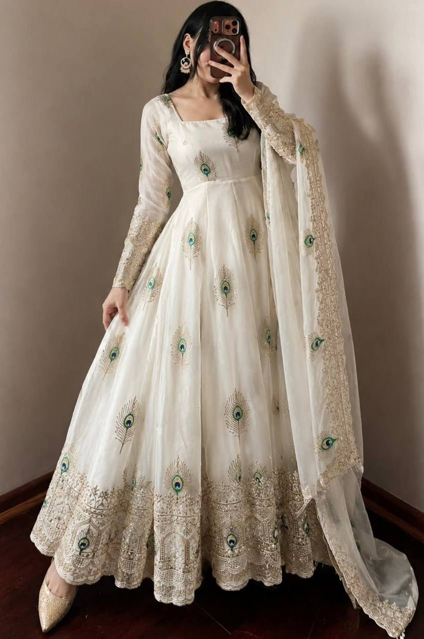 A woman wearing a long white or ivory Anarkali gown with green and gold peacock feather embroidery and a matching sheer dupatta, photographed in a mirror selfie against a plain indoor wall.
