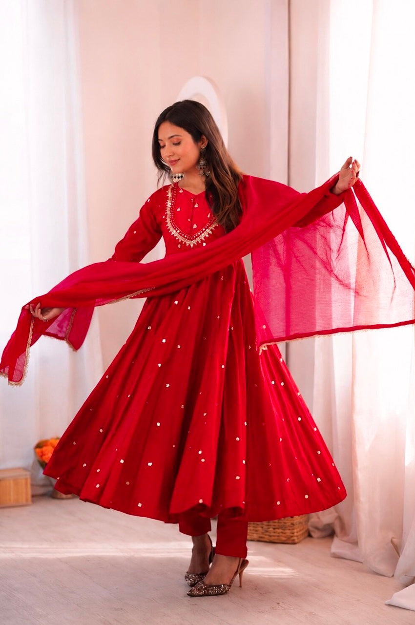 A woman modeling a vibrant red Indian ethnic outfit, featuring a long-sleeved anarkali-style dress with silver mirror work embellishments, matching trousers, and a sheer red dupatta, set against a soft beige background with white curtains.