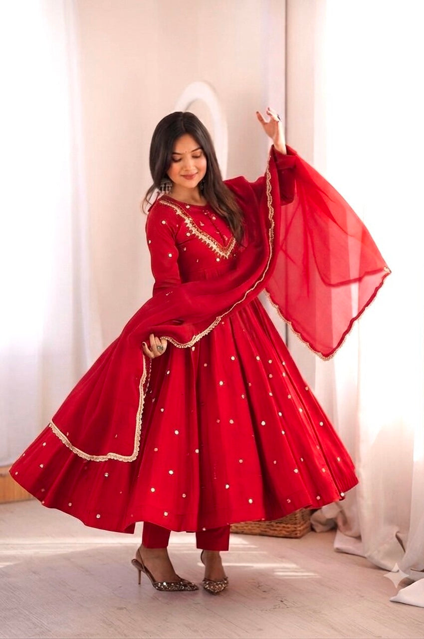 A woman modeling a vibrant red Indian ethnic outfit, featuring a flared red anarkali-style skirt with gold embroidered borders and small gold dots, a matching red kurta with gold embroidery, and a sheer red dupatta, set against a soft, warm neutral background.