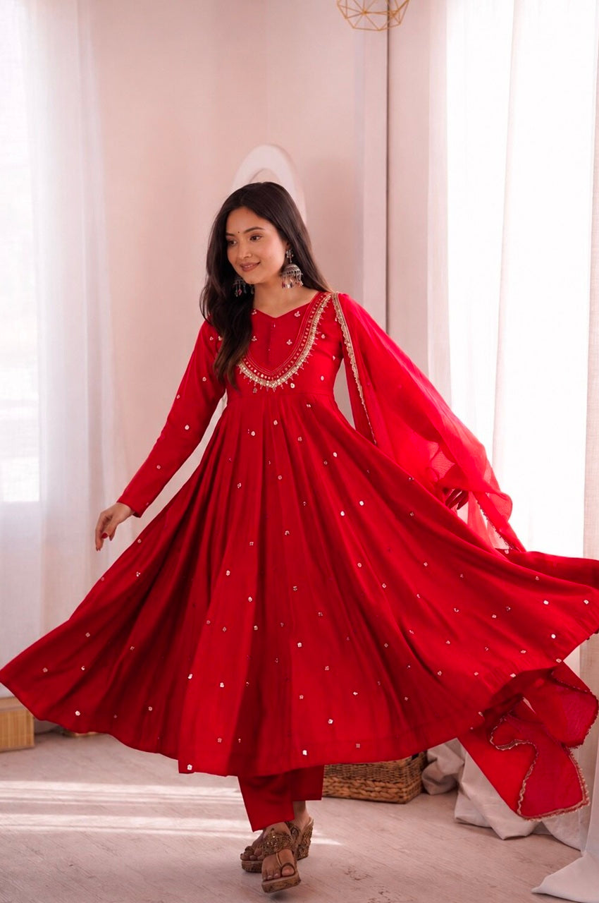 A woman wearing a bright red traditional Anarkali gown with silver mirror work embroidery, captured mid-twist to show the full flowing skirt and dupatta against a soft pink interior background.