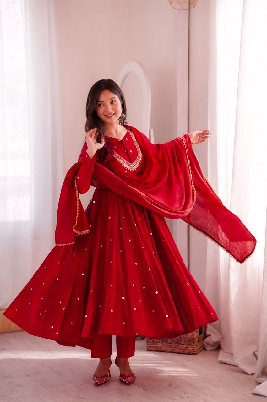 A woman modeling a vibrant red traditional Indian outfit with a flared skirt, matching pants, and a sheer dupatta with gold embroidery, set against a soft, light-colored room with white curtains.
