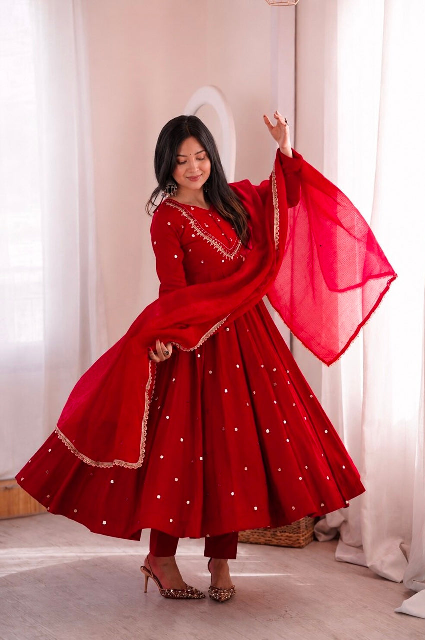 A woman wearing a vibrant red traditional Indian outfit with a matching dupatta, featuring gold embroidery and silver mirror work, posing in a softly lit room with white curtains.