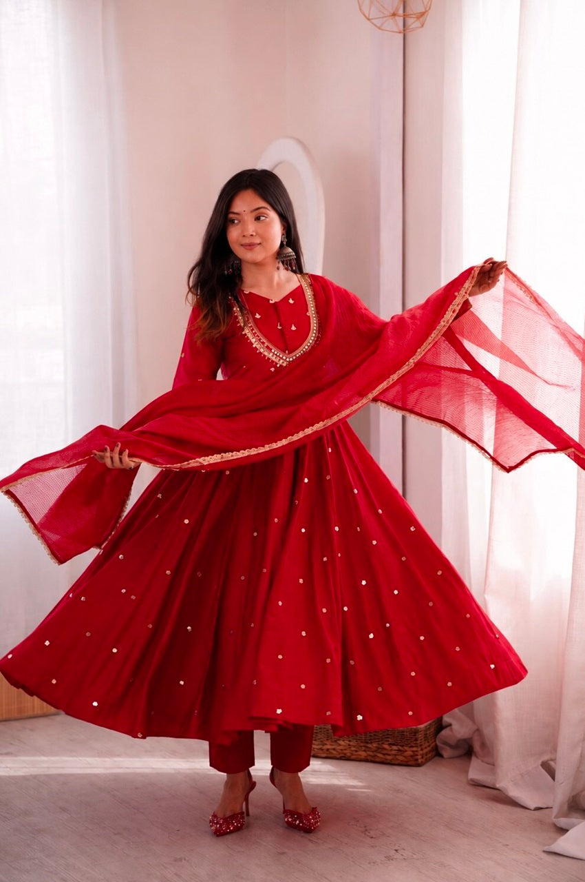 A woman wearing a vibrant red traditional Indian outfit with a matching dupatta, decorated with small gold embellishments, posing in a bright room with white curtains.