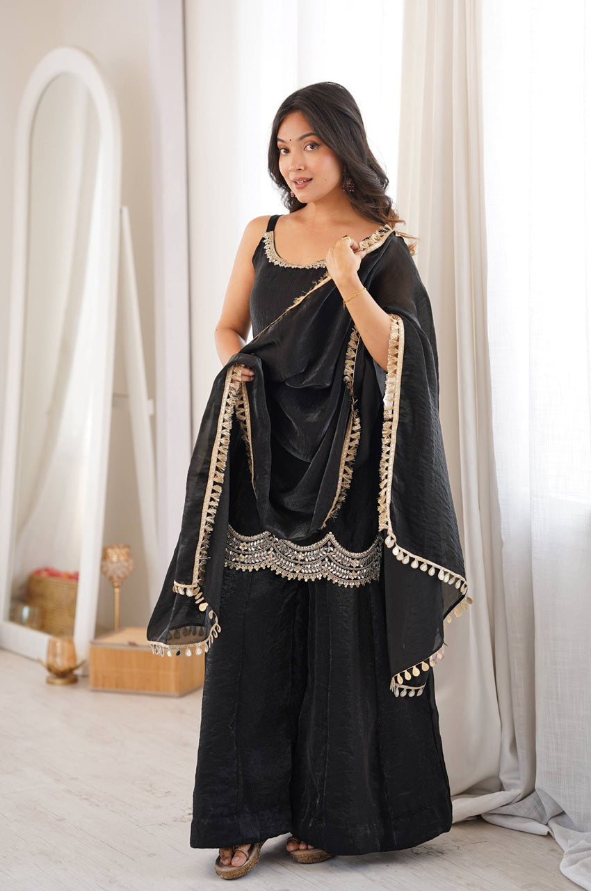 A woman modeling a black traditional outfit with gold embroidered borders, consisting of a sleeveless top, wide-leg pants, and a draped scarf, in a bright indoor setting with white curtains.