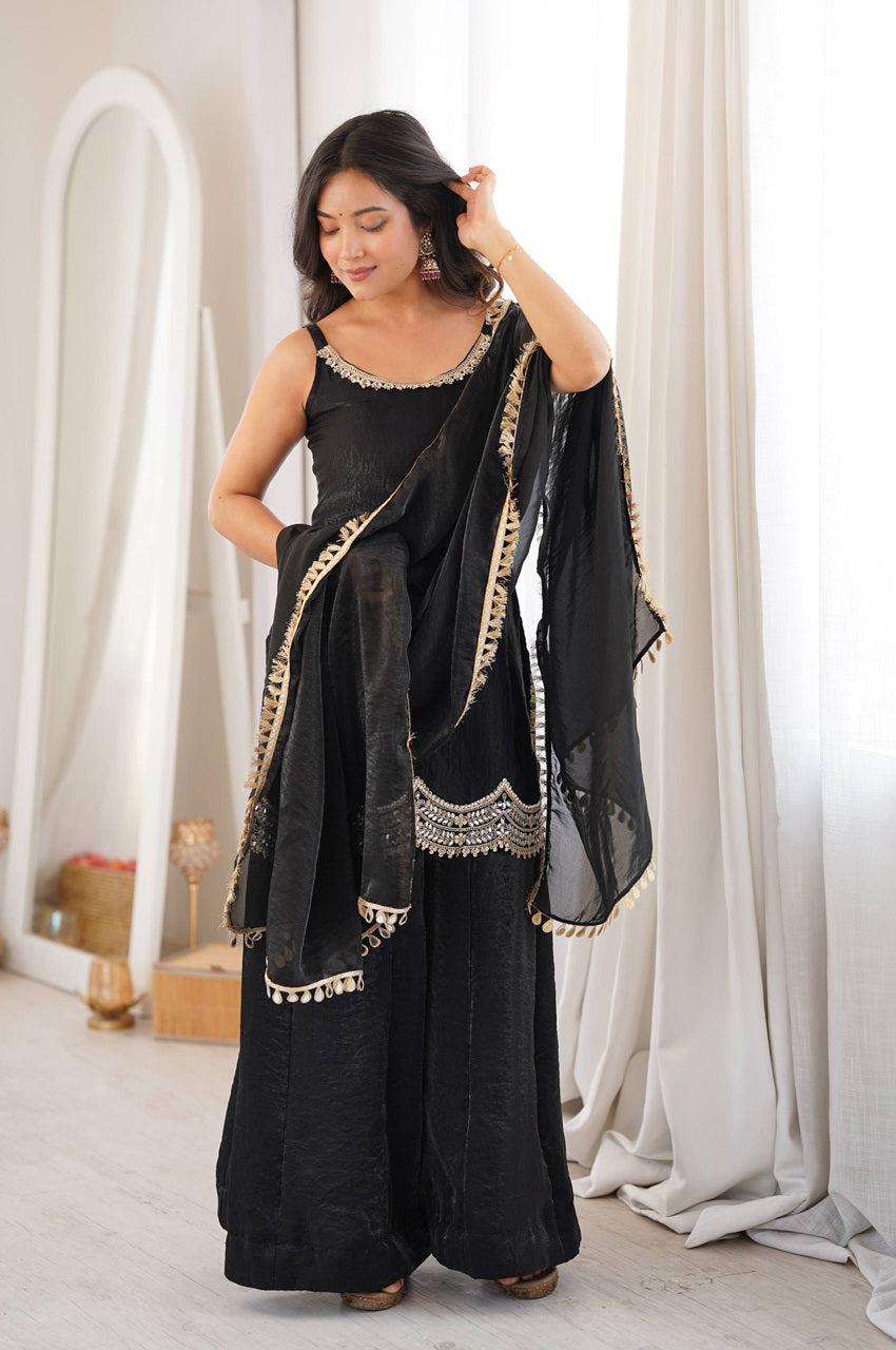 A woman modeling a black traditional outfit with a matching dupatta in a bright, minimalist room with white curtains and a mirror.