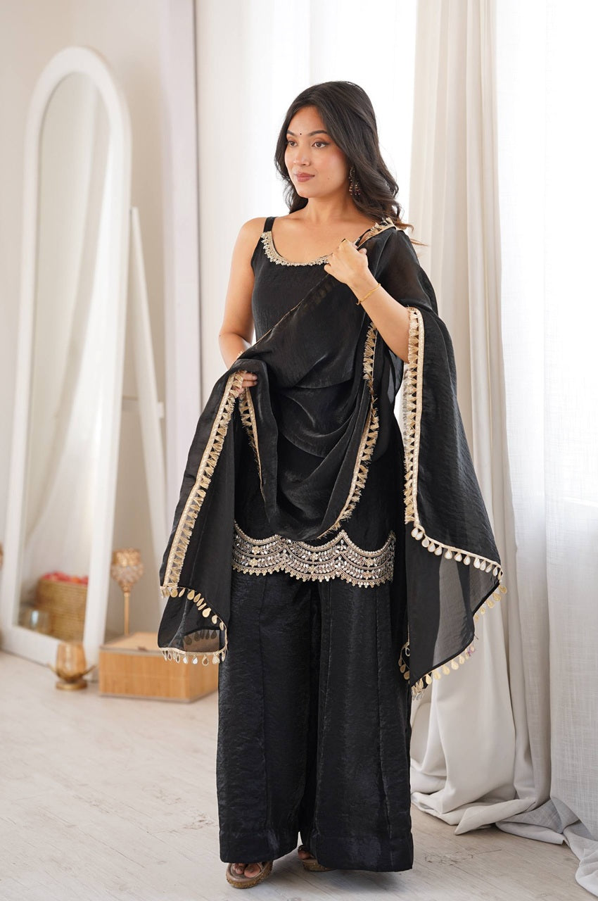 A woman models a black three-piece traditional outfit with beige embroidered borders and tassels, consisting of a sleeveless top, wide-leg pants, and a draped dupatta, in a bright, modern room with white curtains.