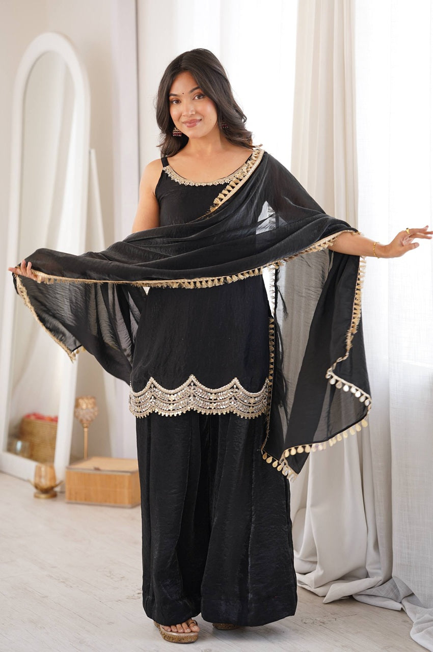 A woman modeling a black three-piece salwar kameez set with gold embroidery, featuring a sleeveless tunic, wide-leg pants, and a matching dupatta draped over her shoulders in a bright indoor setting.