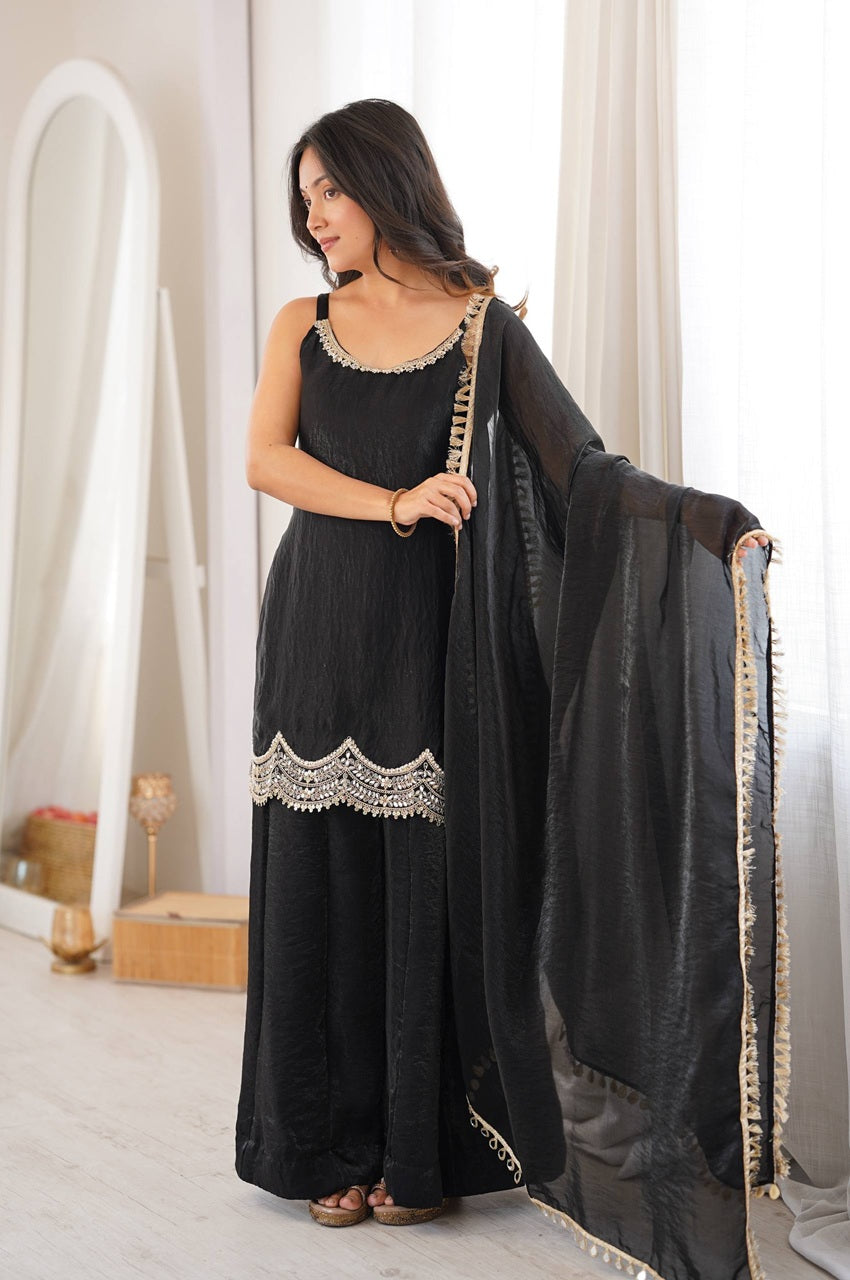 A woman modeling a black traditional outfit with silver embroidered details, featuring a sleeveless top, wide-leg pants, and a matching sheer dupatta draped over her arm in a bright indoor setting.