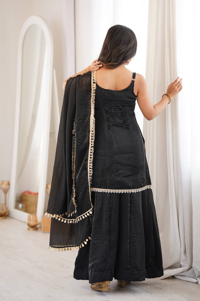 A woman viewed from behind wearing a black traditional outfit with a matching draped scarf, standing in a bright indoor room with white curtains and a mirror in the background.